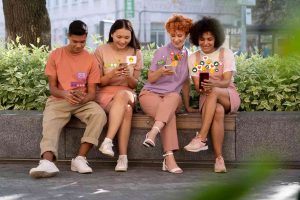 A group of young adults sitting on a bench outdoors, smiling while interacting with their smartphones and surrounded by digital icons, representing the hyper-connected lifestyle and digital habits of Generation Z.
