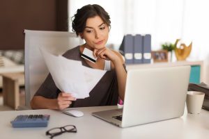A thoughtful woman sitting at a desk, reviewing financial documents with a credit card in hand, reflecting on expenses and analyzing her credit limits using a laptop.